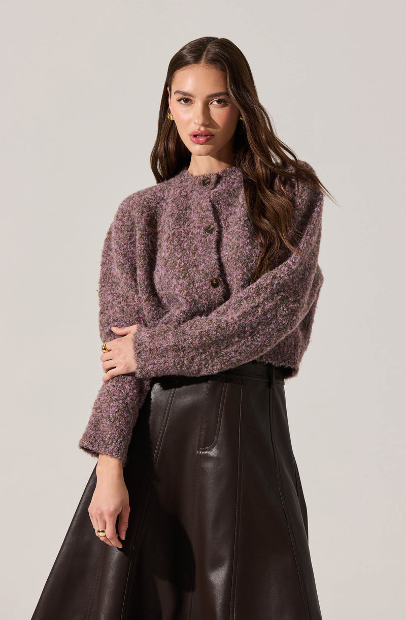A woman is wearing a textured, long-sleeved cardigan in shades of purple and gray with a high neckline and large buttons, paired with high-waisted, loose-fitting black leather pants.