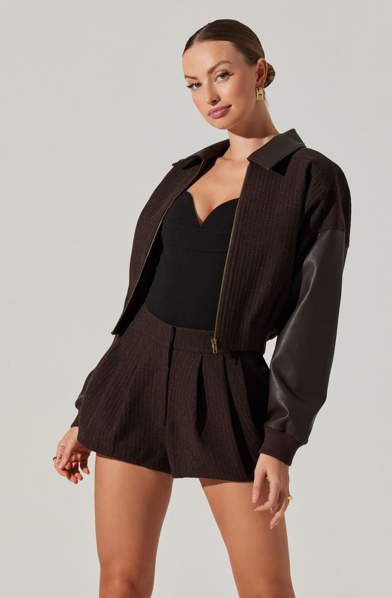 A woman is wearing a dark, high-waisted pleated shorts made of textured fabric, paired with a fitted black top with a sweetheart neckline. Over the top, she has a dark jacket with a ribbed body, leather sleeves, and a front zipper. The jacket features a collar and appears to be made of a combination of fabric and leather materials.