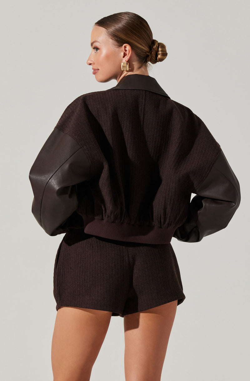 A woman is wearing a black bomber jacket with large, textured sleeves made of leather and knit fabric, featuring a ribbed waistband. She pairs it with matching black knit shorts and has her hair styled in a low, neat bun. She is also wearing statement gold earrings.
