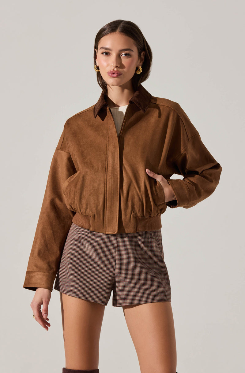 A young woman is wearing a brown, zip-up, suede jacket with a collar and elasticized waistband. She is also dressed in high-waisted, plaid shorts with a subtle checkered pattern in shades of brown and black.