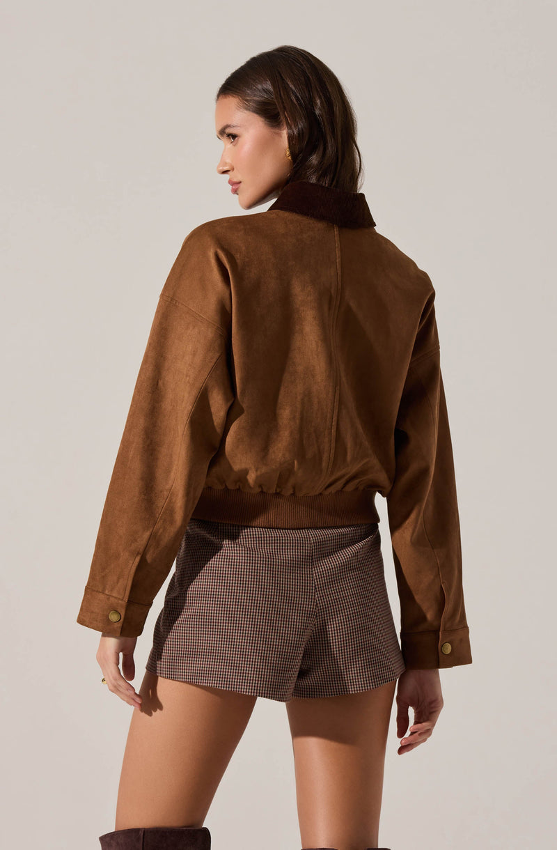 A woman is wearing a brown suede jacket with a darker collar, cargo-style sleeves with button cuffs, and a ribbed hem. She pairs this with high-waisted shorts featuring a small checkered pattern in dark and light brown tones.