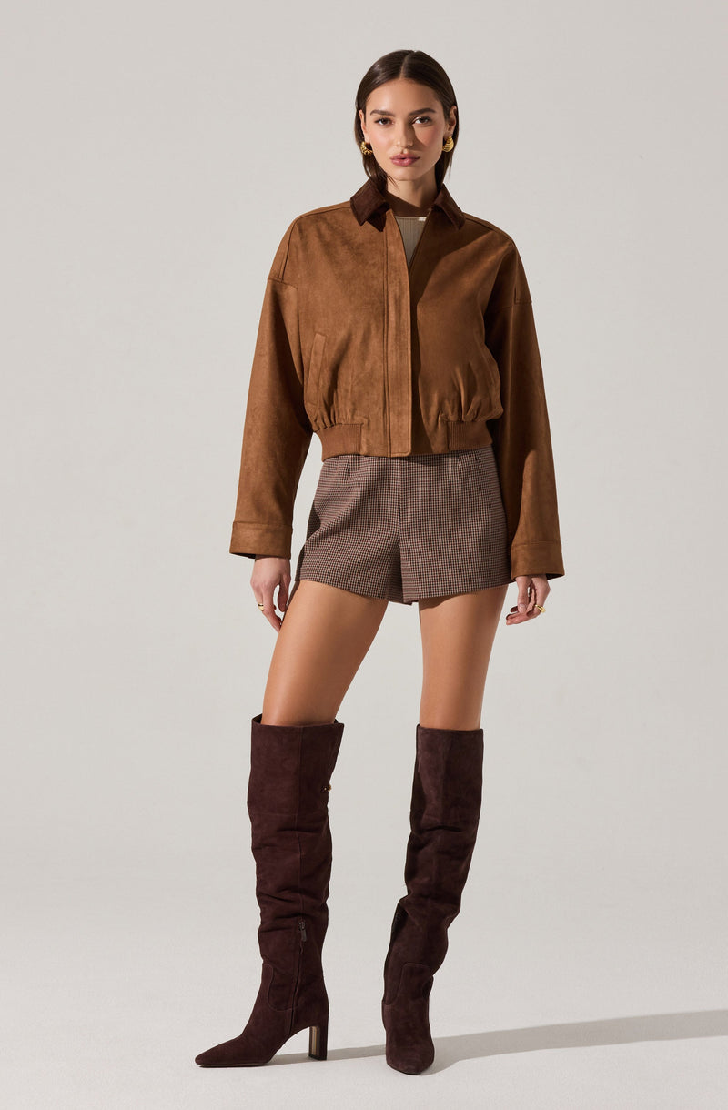 A woman is wearing a brown cropped jacket with a collar and long sleeves. She has on high-waisted shorts with a small checkered pattern. She is also wearing tall, dark brown suede over-the-knee boots with high heels.