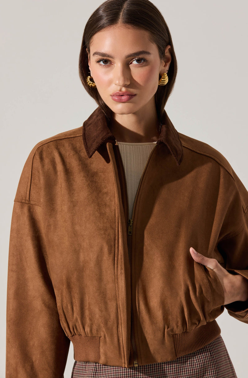 A woman is wearing a brown suede jacket with a collar, zip closure, and ribbed cuffs at the sleeves. She has paired it with gold hoop earrings and a beige top underneath.
