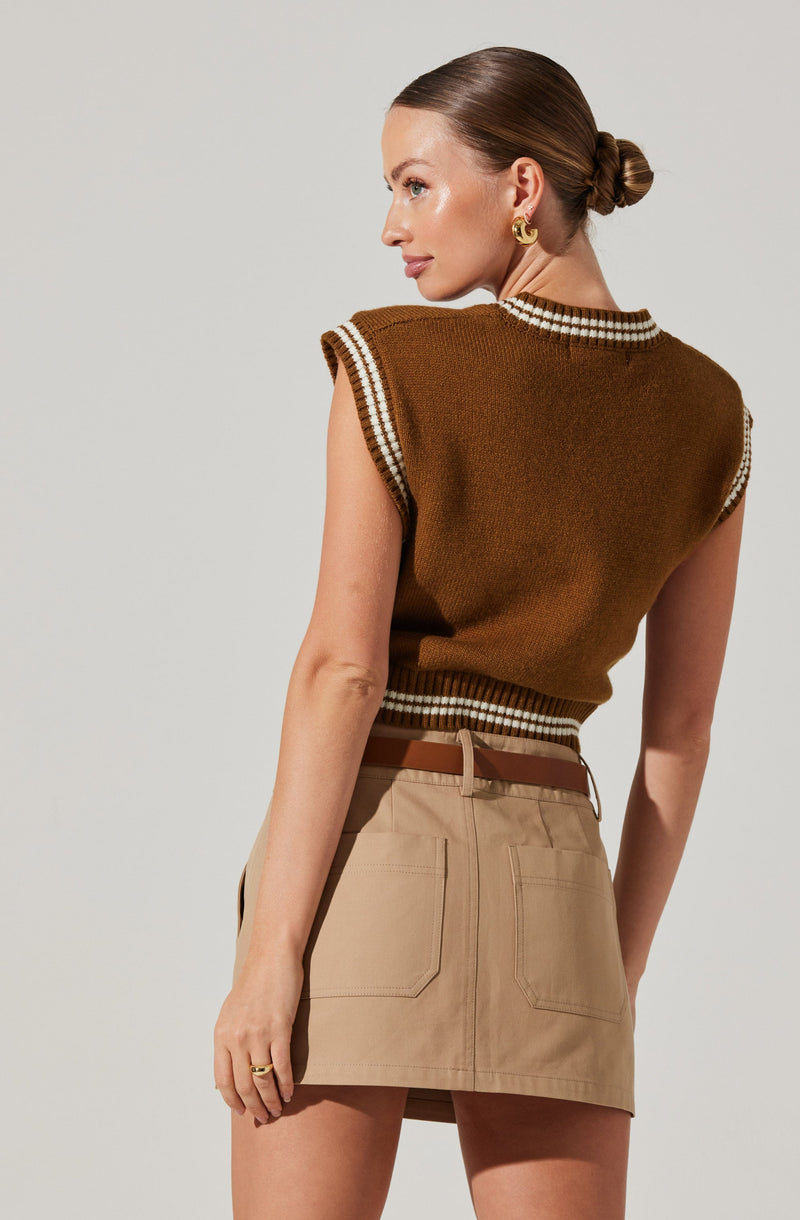 A woman is wearing a sleeveless, brown knitted sweater with white and brown striped trim around the collar, armholes, and waistband. She also has on a beige skirt with large back pockets and a brown belt. She has gold hoop earrings and a gold ring. Her hair is styled in a low, twisted bun.