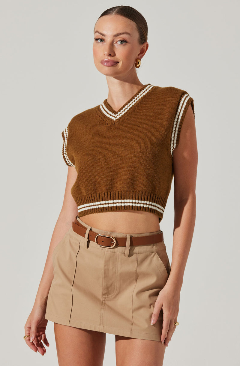 A woman is wearing a sleeveless brown knitted vest with white and brown striped trim around the V-neckline, armholes, and hem. She pairs it with a beige high-waisted skirt with a front zipper and seam details, secured with a brown belt with a gold buckle.