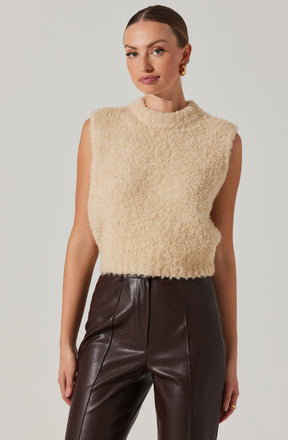 A woman is wearing a sleeveless, beige, textured, high-neck sweater that appears soft and cozy. She has paired it with high-waisted, dark brown leather pants with vertical seam detailing. She is accessorized with gold earrings and a ring on her right hand.