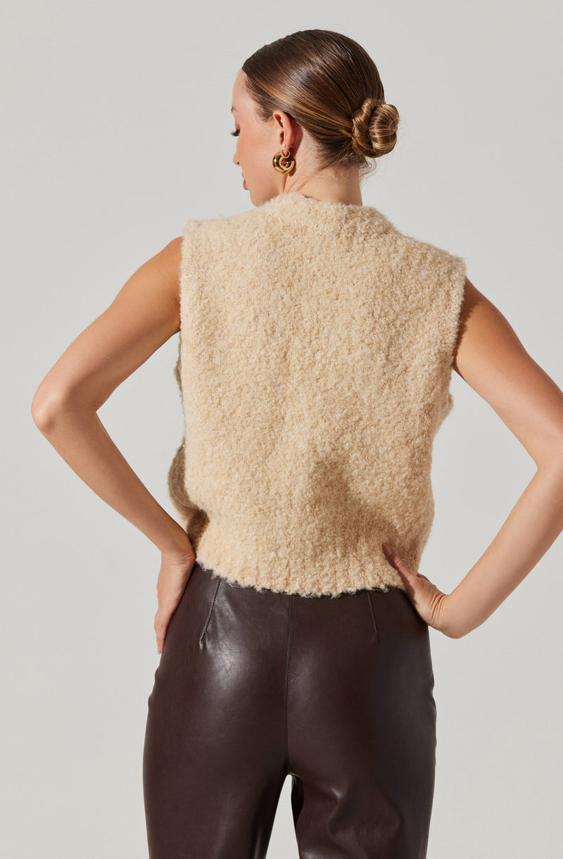 A woman is wearing a sleeveless, beige, fuzzy textured vest that has a slightly boxy fit. She pairs it with high-waisted, dark brown leather pants.