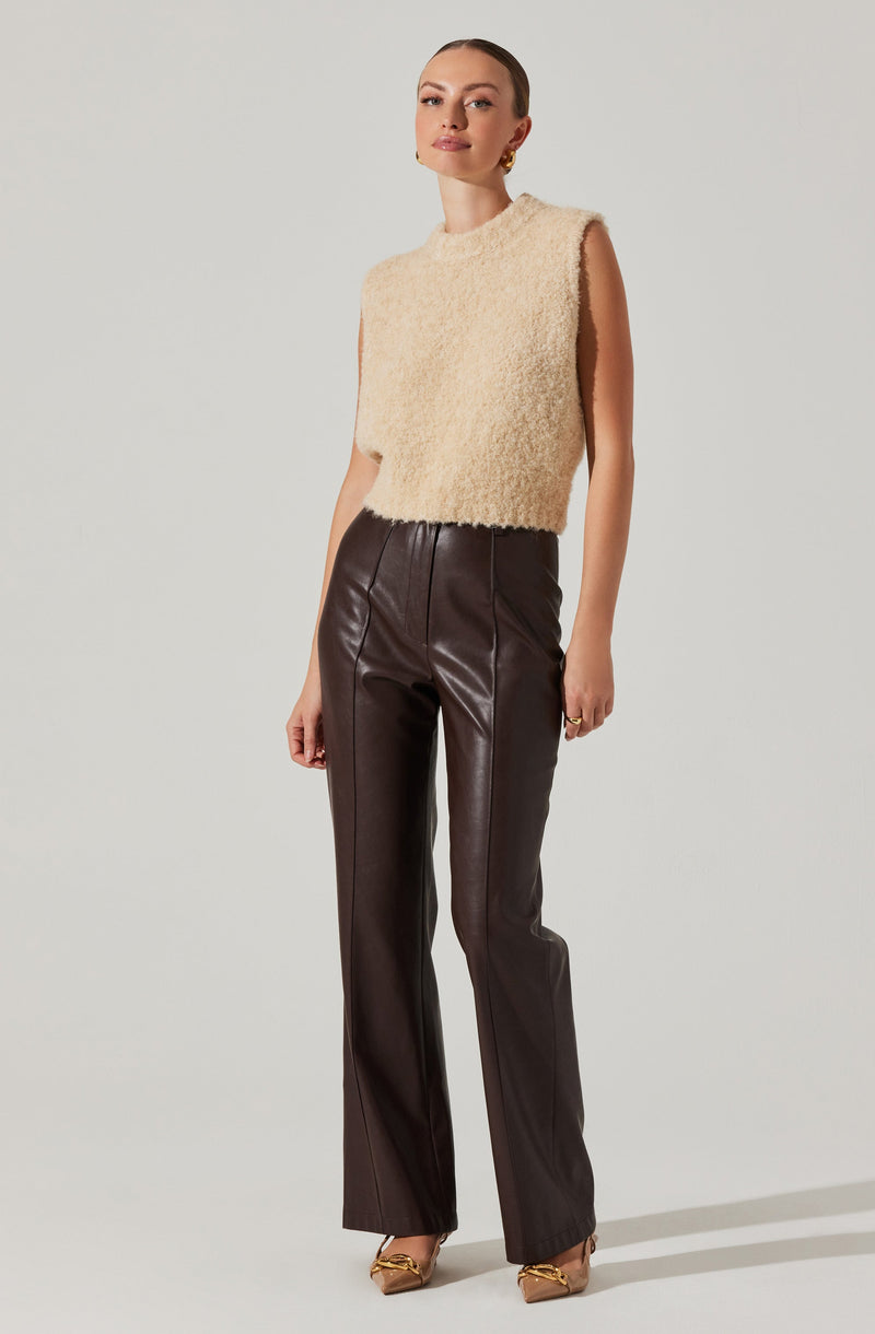A woman is wearing a sleeveless, high-neck, fuzzy beige top paired with high-waisted, dark brown leather pants that have a straight-leg fit. She is accessorized with gold earrings and a ring, and is wearing pointed-toe heels with a metallic detail.