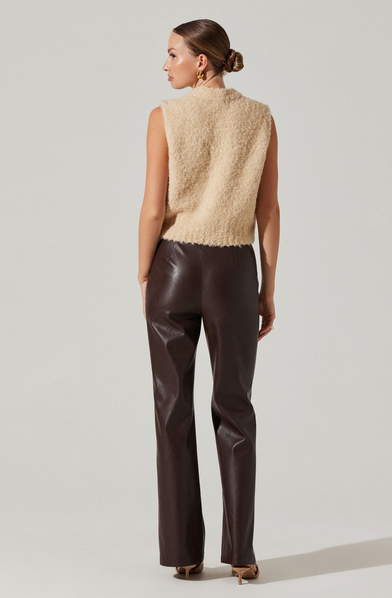 A woman is wearing a sleeveless, beige, fuzzy vest made of a soft, textured material. She pairs it with high-waisted, dark brown leather pants that fit snugly through the hips and thighs, and have a straight-leg cut. She is also wearing beige high-heeled shoes. Her hair is styled in a neat low bun, and she has gold hoop earrings.