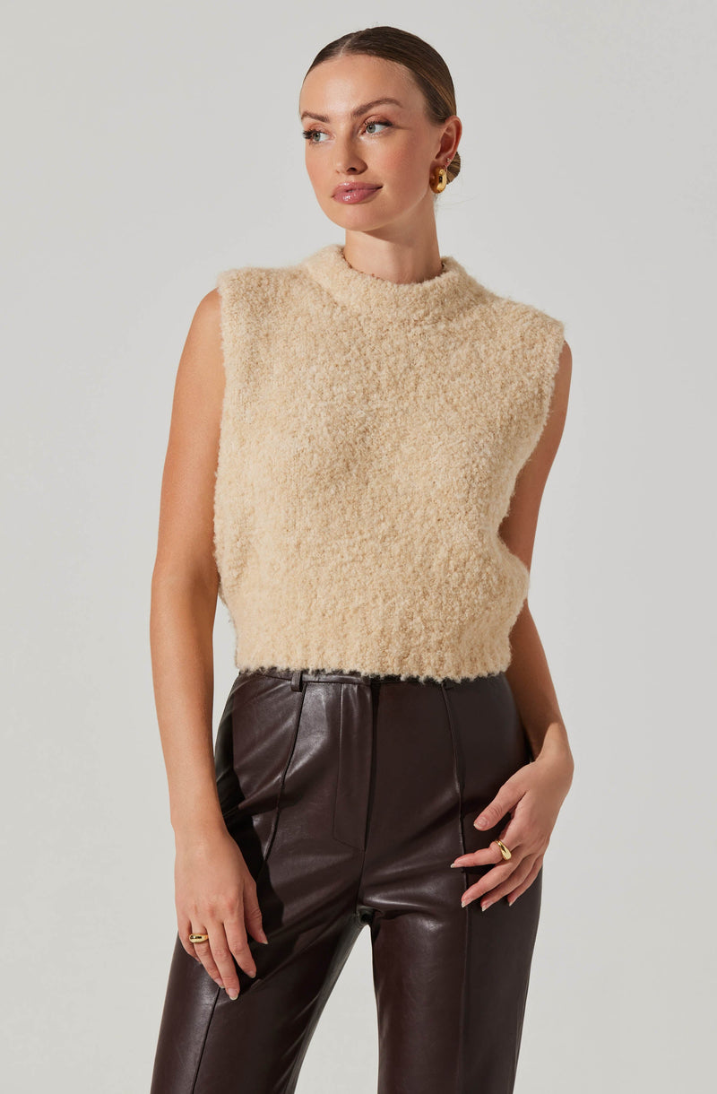A woman is wearing a sleeveless, high-neck, cream-colored, fuzzy sweater paired with high-waisted, dark brown leather pants.