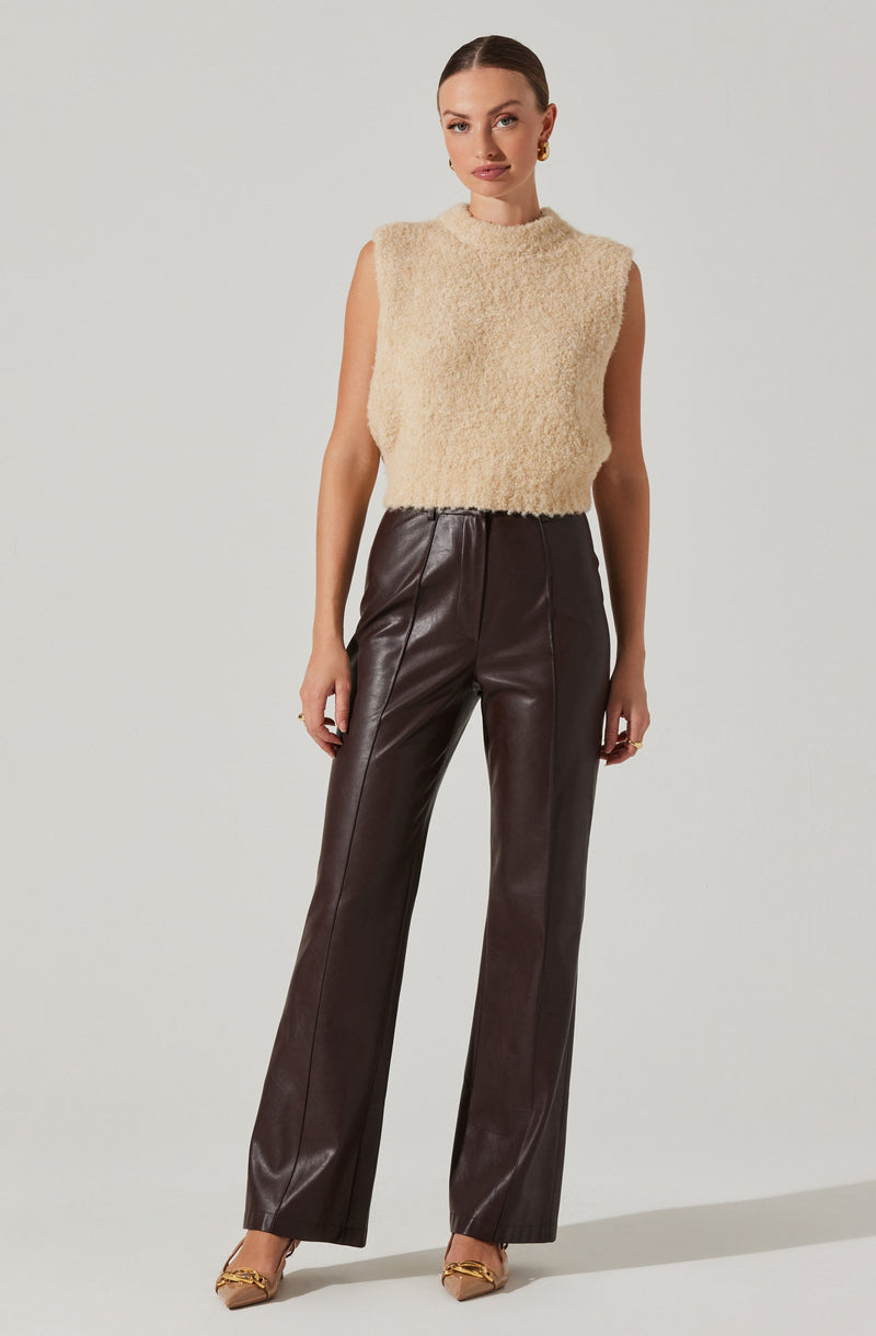 A woman is wearing a sleeveless, beige, fuzzy knit top with a high neckline. She pairs it with high-waisted, dark brown leather pants that have a straight-leg fit. She completes her outfit with beige high-heeled shoes featuring a chain detail on the front. Her hair is slicked back, and she has gold earrings.
