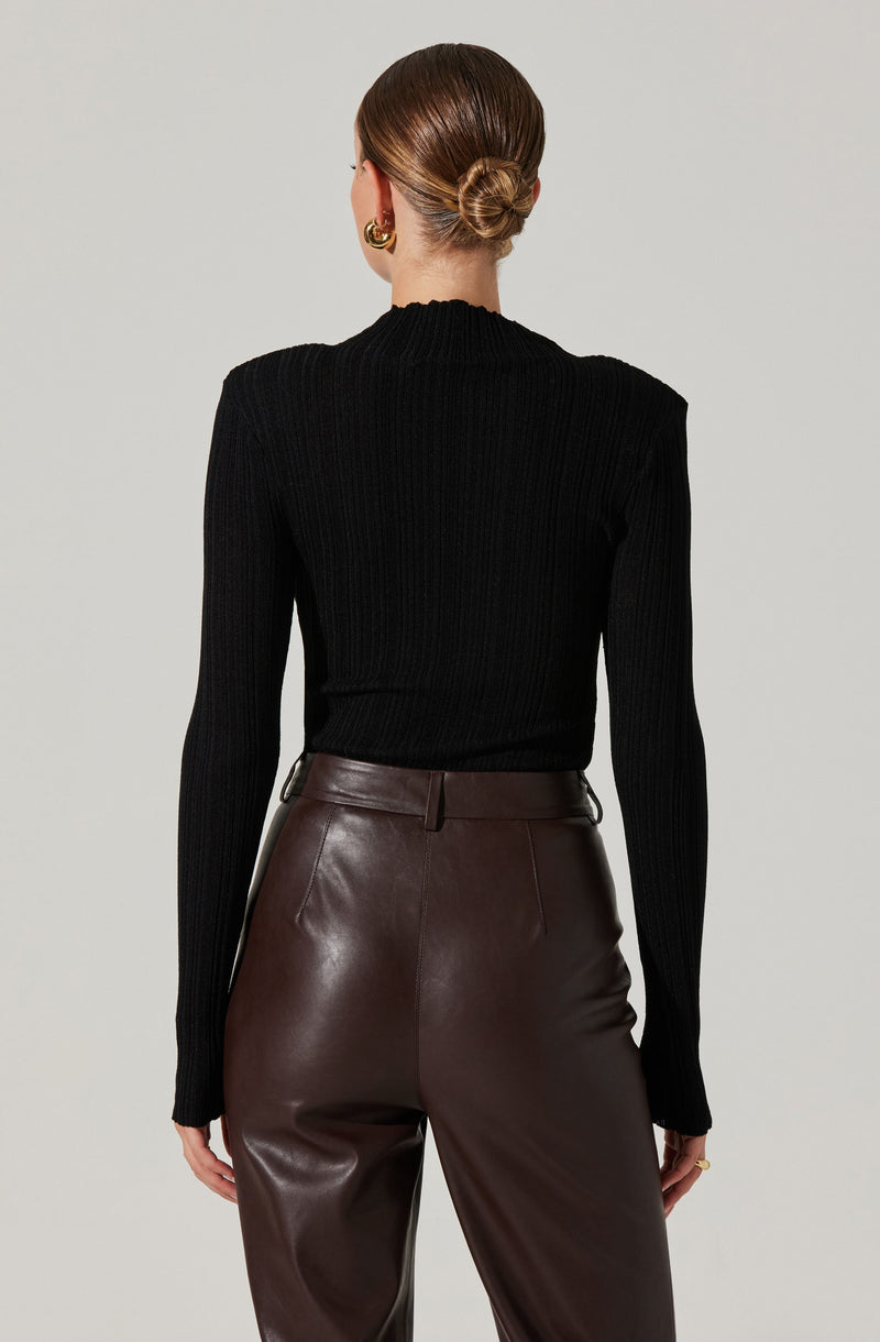 A woman is wearing a fitted black ribbed sweater with a high neckline and long sleeves. She pairs it with high-waisted, dark brown leather pants that have a smooth finish and a waistband with belt loops. Her hair is styled in a low, neat bun, and she is accessorized with large, gold hoop earrings.