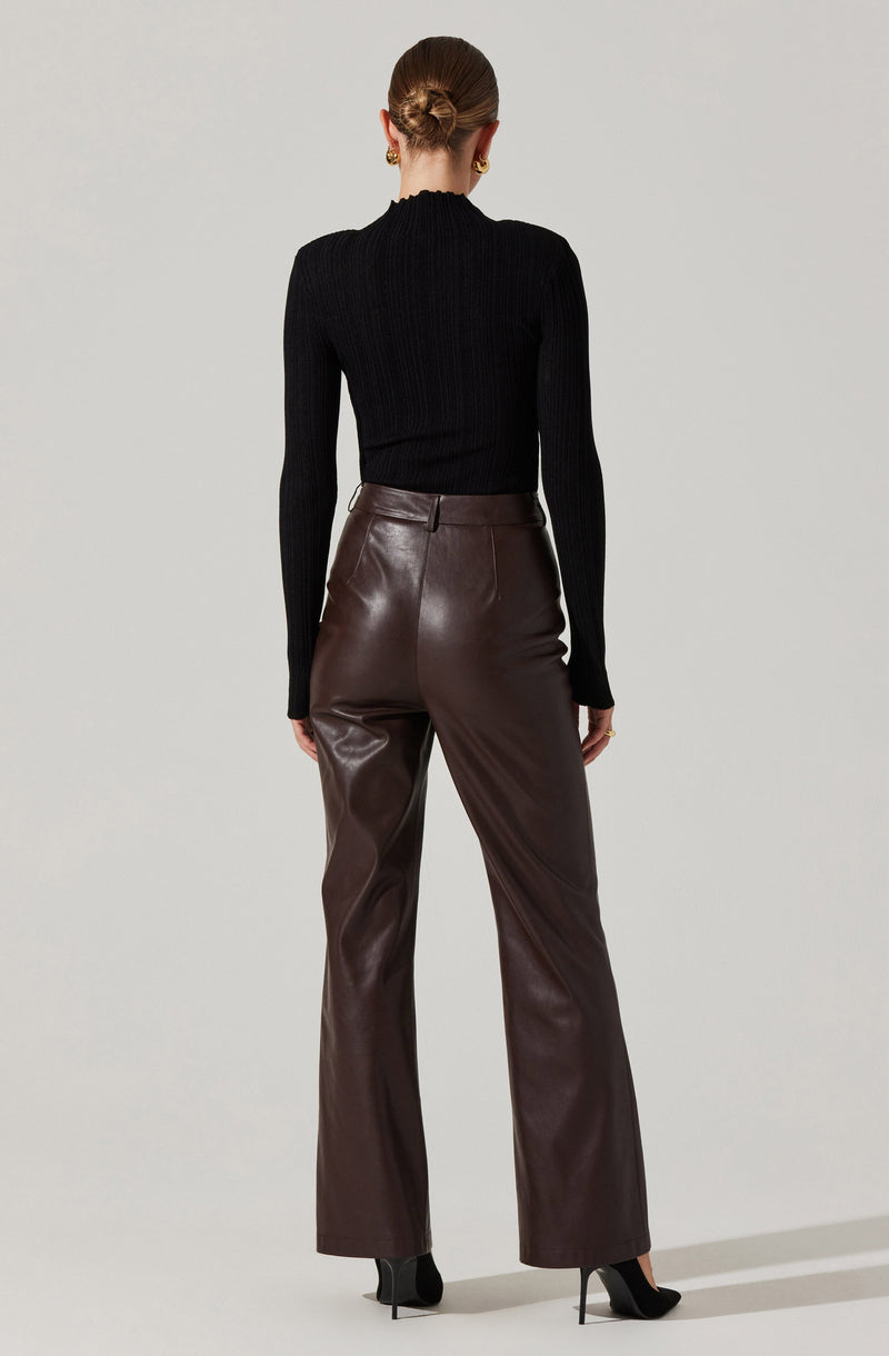 A person is wearing a fitted, long-sleeve black ribbed turtleneck sweater paired with high-waisted, wide-leg brown leather pants. They are also wearing black pointed high-heeled shoes and gold earrings. Their hair is styled in a neat bun.