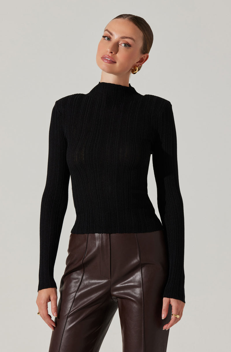 A woman is wearing a fitted black ribbed turtleneck sweater with long sleeves and a high neckline. She is also wearing high-waisted brown leather pants with visible vertical seams.