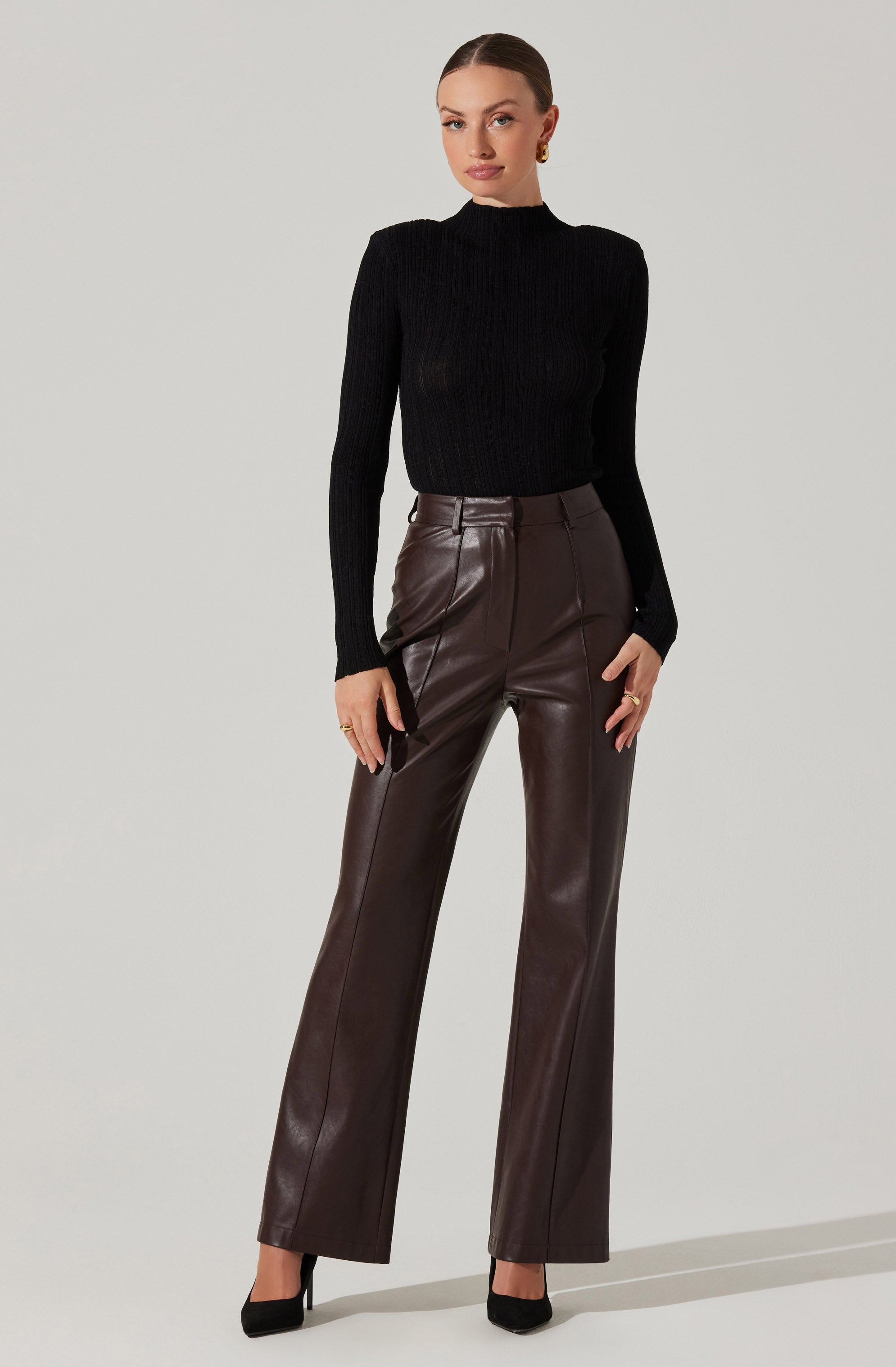 A woman is wearing a fitted black ribbed long-sleeve turtleneck sweater and high-waisted, wide-leg brown leather pants. She is also wearing black high-heeled shoes.