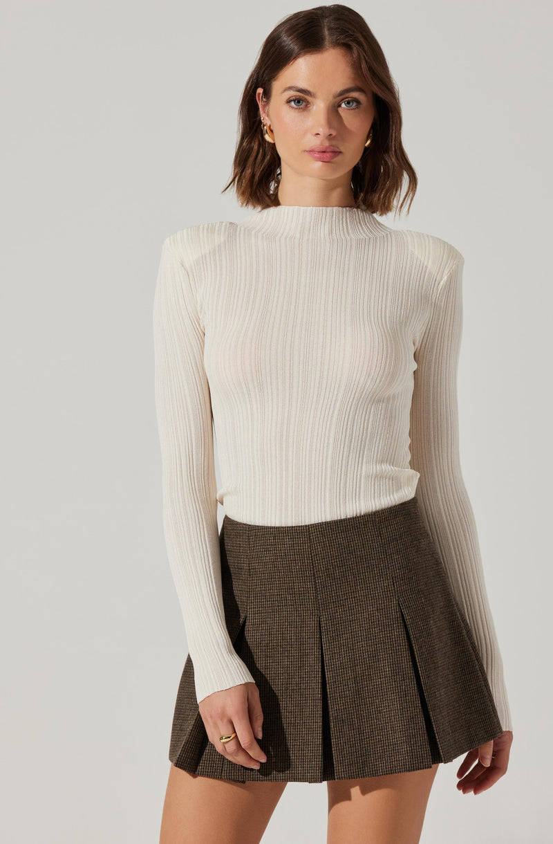 A woman is wearing a long-sleeved, cream-colored ribbed sweater with a high neckline, paired with a brown plaid pleated skirt.