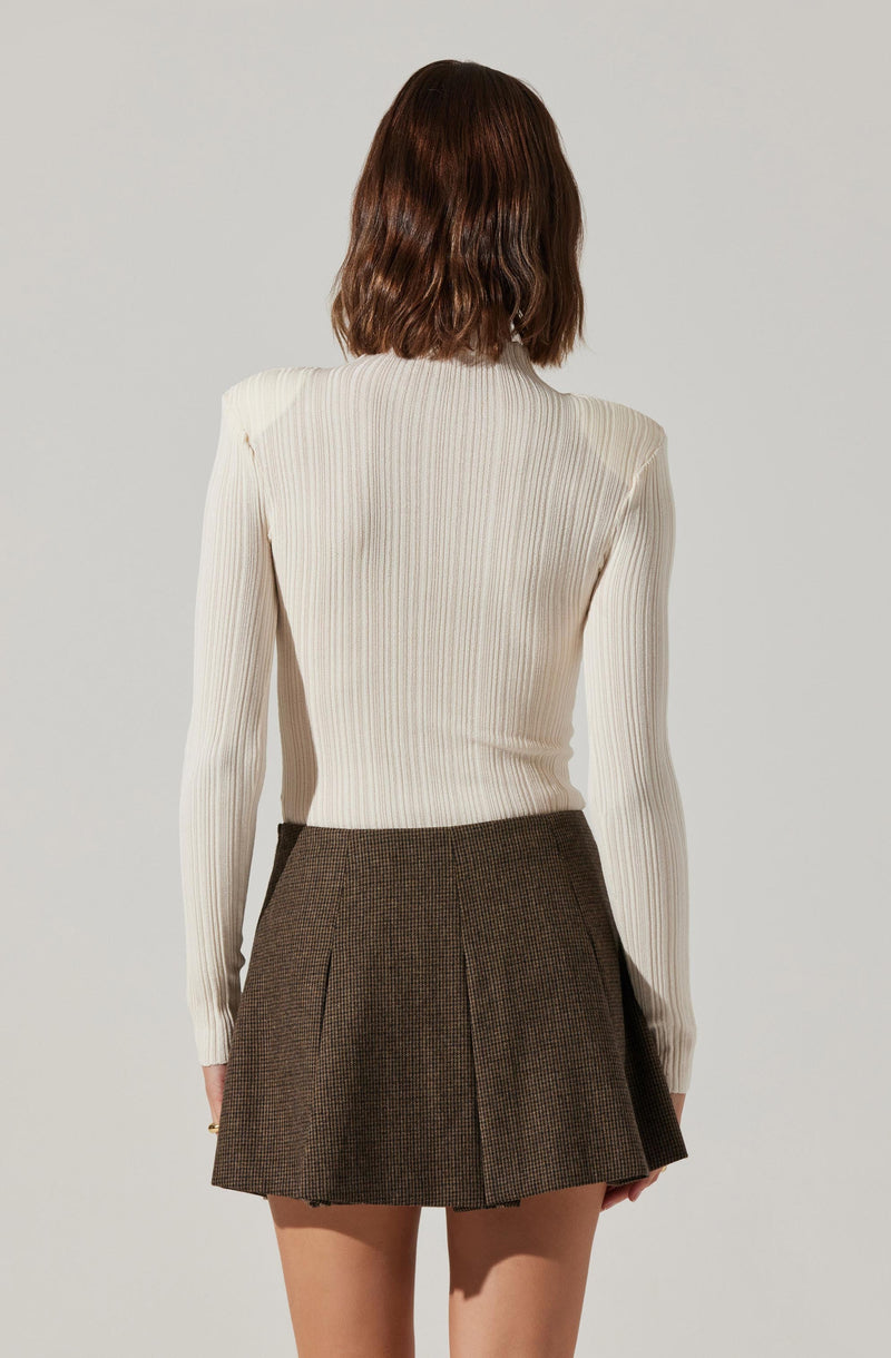A woman is wearing a fitted, long-sleeve, ribbed cream-colored sweater and a high-waisted, A-line brown plaid skirt with pleats.