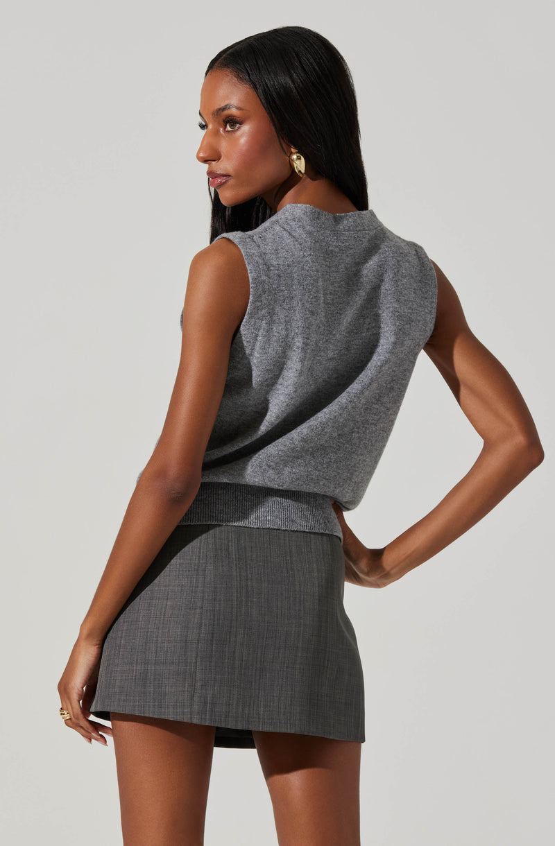 A woman is wearing a sleeveless, grey knitted top with a relaxed fit and a high neckline, paired with a grey plaid mini skirt. She has long, straight black hair and is accessorized with gold earrings.