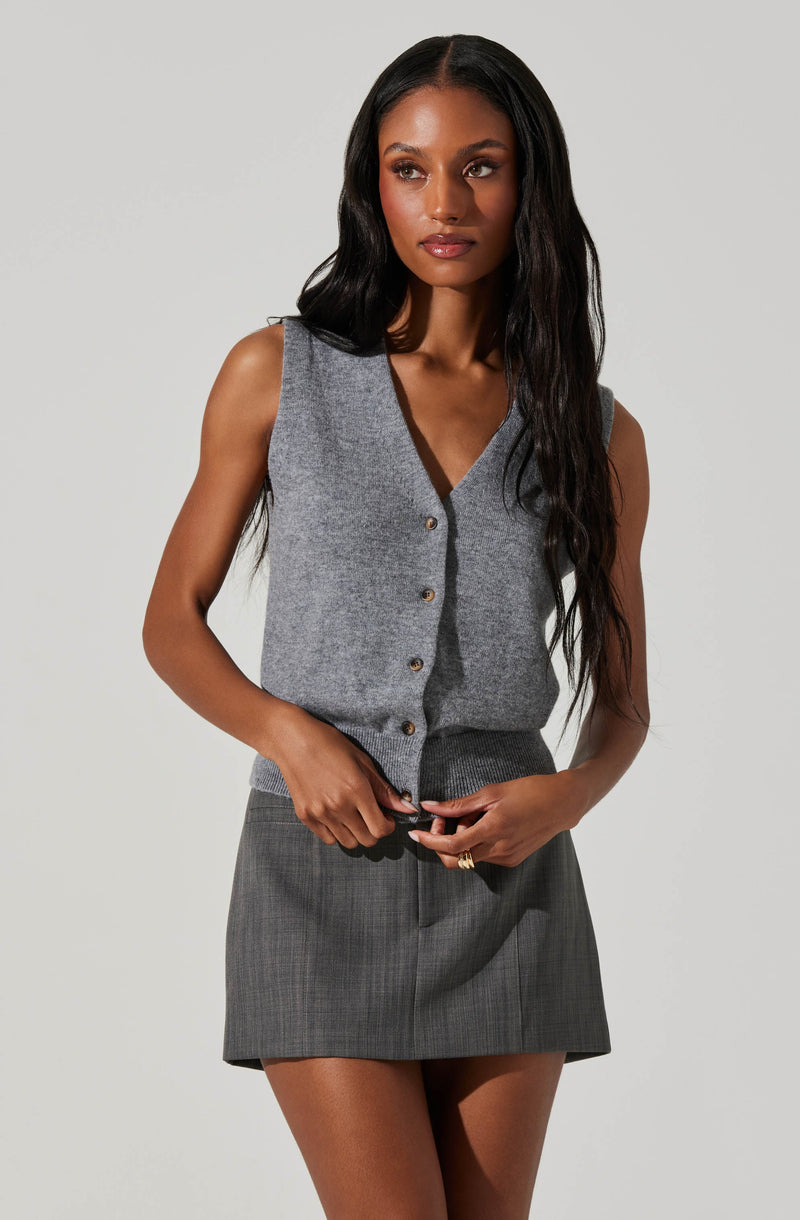 A woman is wearing a sleeveless, V-neck gray vest with buttons down the front. The vest appears to be made of a soft, knit fabric. She is also wearing a high-waisted gray plaid skirt that is above knee length.