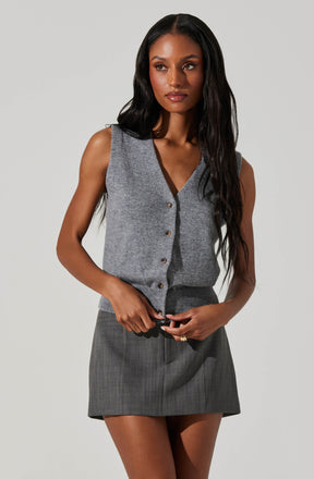 A woman is wearing a sleeveless, V-neck gray vest with buttons down the front. The vest appears to be made of a soft, knit fabric. She is also wearing a high-waisted gray plaid skirt that is above knee length.