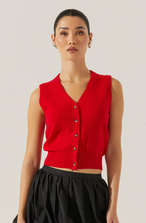 A woman is wearing a sleeveless red knit vest with a V-neckline and button-down front. The vest has a fitted ribbed hem at the waist, creating a cropped look. She is paired with black, loose, gathered pants.