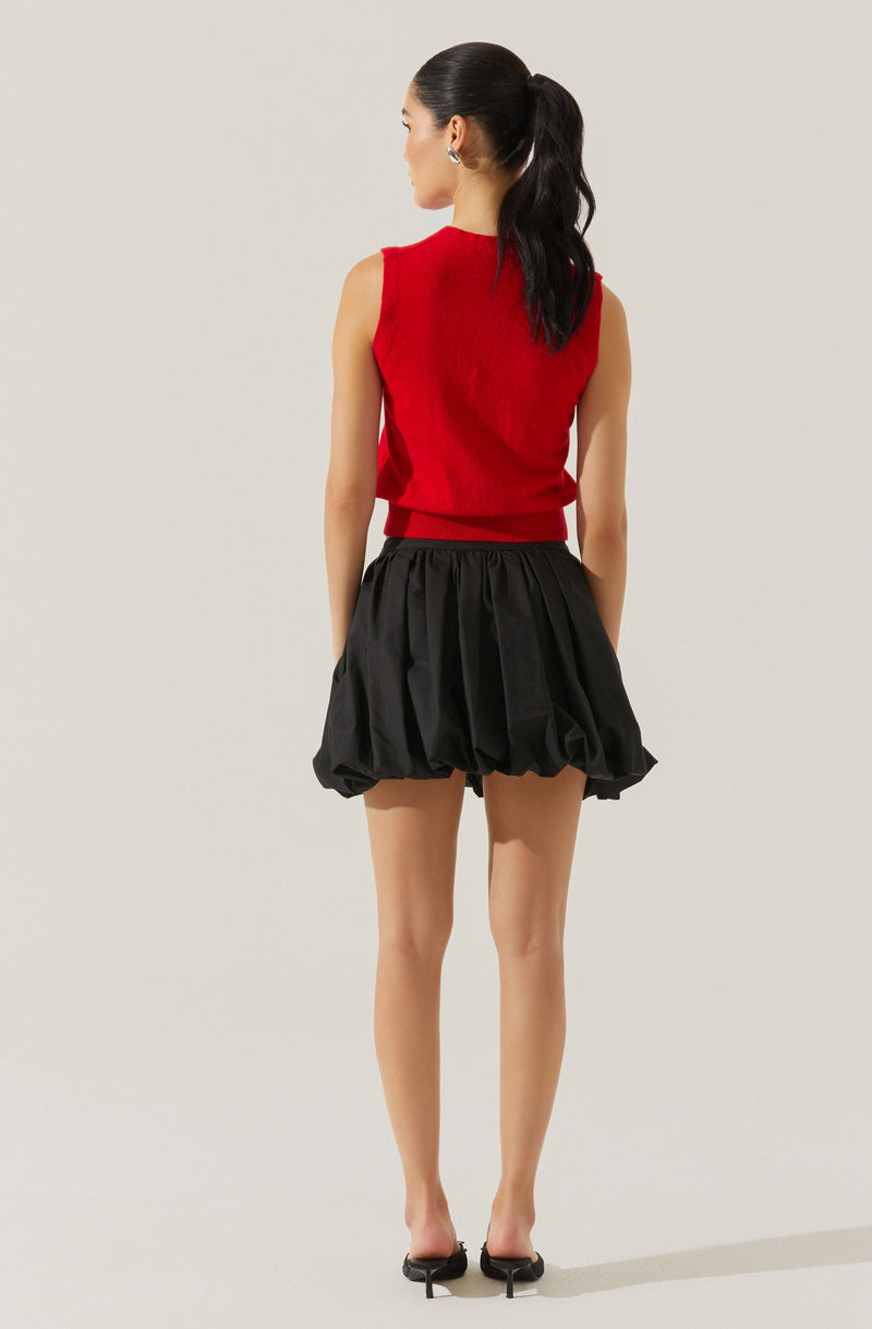 A woman is wearing a sleeveless, red knit top with a ribbed hem, paired with black, voluminous, bubble shorts that have an elastic waistband and a gathered, puffed appearance. She has her hair tied back in a high ponytail and is wearing black high heels.