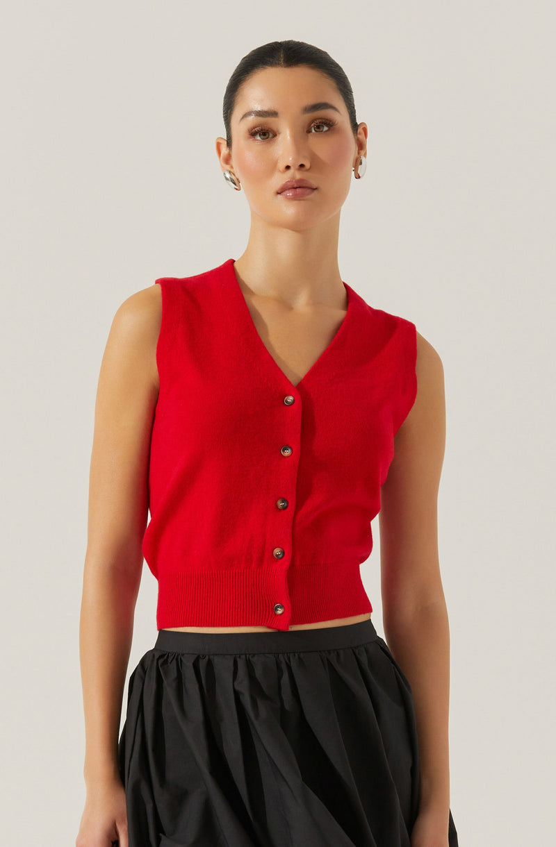 A woman is wearing a sleeveless red knit vest with a V-neckline and a row of metallic buttons down the front. The vest is fitted at the waist, ending just above the waistline. She pairs it with high-waisted, loose-fitting black pants that have a gathered, slightly puffy appearance.