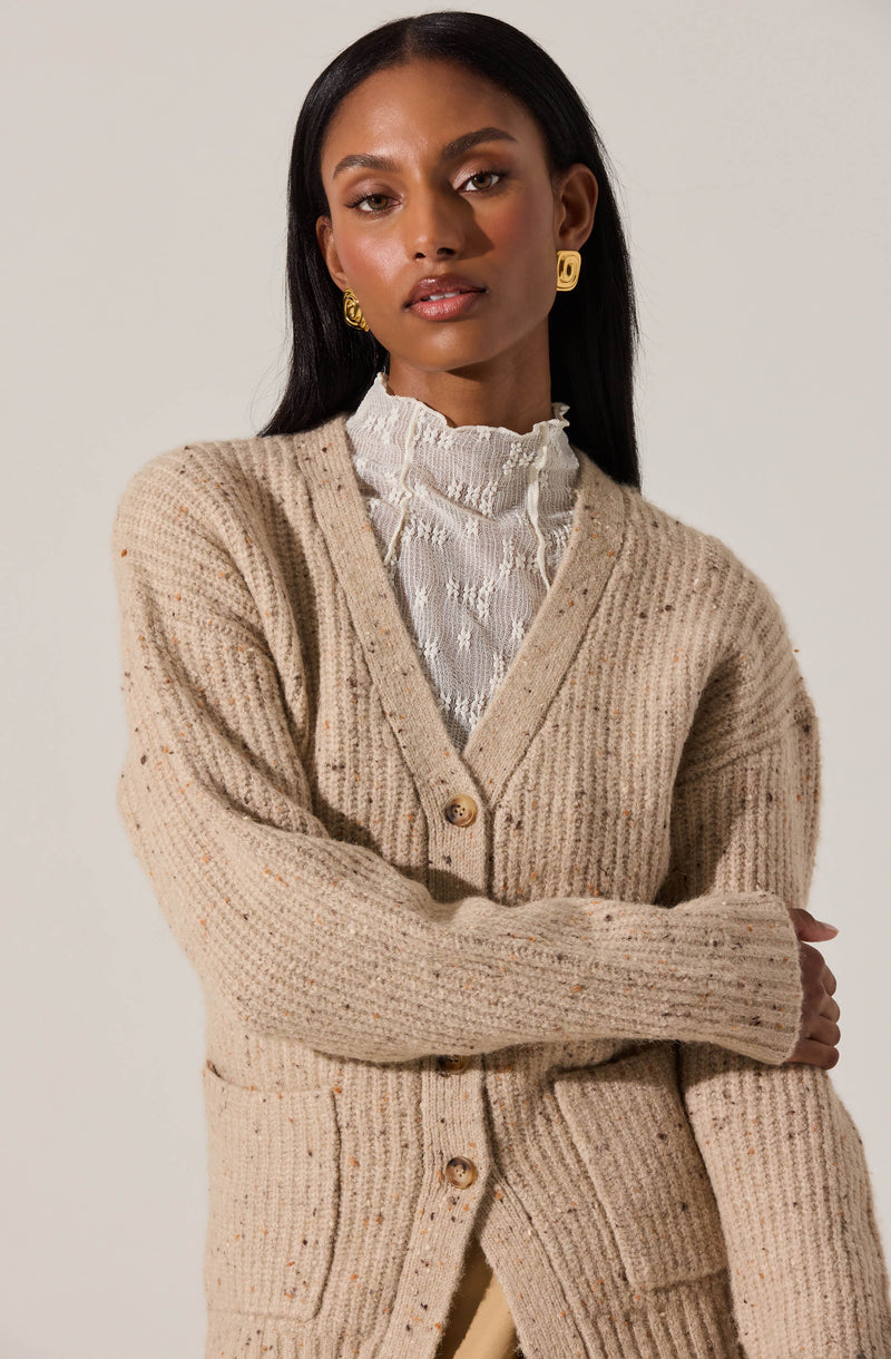 A woman is wearing a beige, chunky knit cardigan with large buttons down the front. Underneath, she has on a white, high-neck blouse with ruffled detailing at the collar. She accessorizes with gold, geometric-shaped earrings.