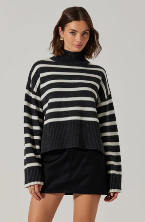 A woman is wearing a black and white horizontally striped turtleneck sweater with a chunky knit design. The sweater features wider black and white stripes and a ribbed hem and cuffs. She pairs the sweater with a black leather mini skirt.