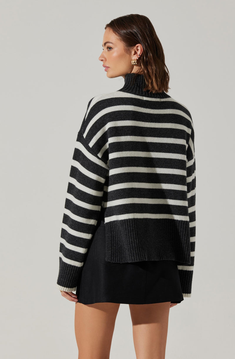 A woman is wearing a loose-fitting, long-sleeved sweater with wide horizontal black and white stripes, a high turtleneck collar, and ribbed cuffs and hem. She pairs it with a black skirt.