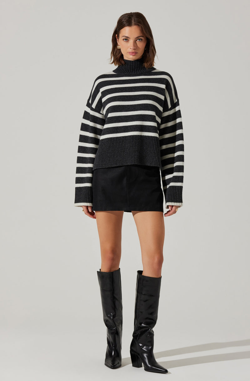 A woman is wearing a black and gray striped turtleneck sweater with oversized sleeves, paired with a black mini skirt and knee-high black shiny leather boots with pointed toes and chunky heels.