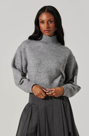 A woman is wearing a chunky, light gray knit sweater with a high turtleneck and long, slightly puffed sleeves. She pairs it with a high-waisted, dark gray pleated skirt.