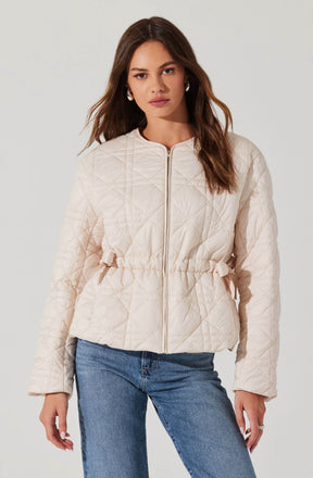A woman is wearing a light-colored, quilted jacket with a front zipper. The jacket features a drawstring waist and a subtle diamond pattern in the quilting design. She has paired it with blue jeans.