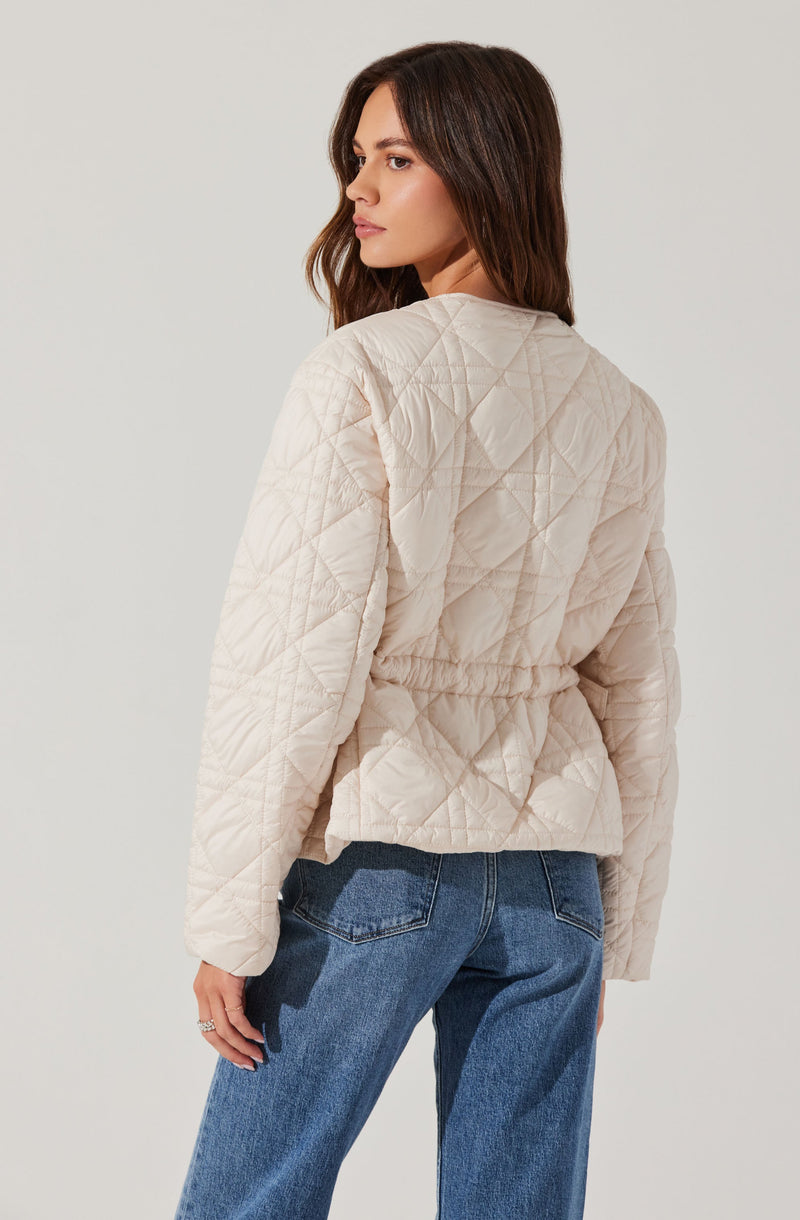 A woman is wearing a light beige quilted jacket with a subtle diamond pattern. The jacket has a gathered waistband and slightly puffed sleeves. She is also wearing medium wash blue jeans.