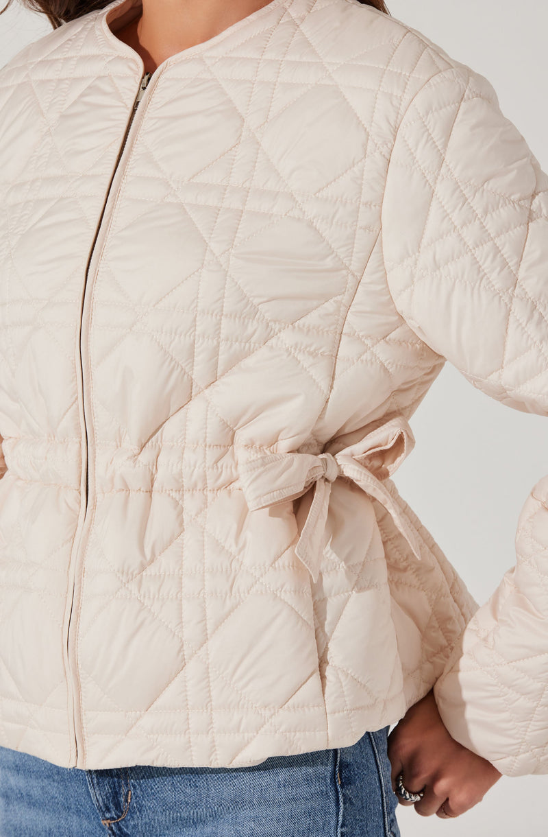 A quilted, cream-colored jacket with a round neckline and a front zipper. The jacket features a subtle diamond pattern, and it has a side tie detail at the waist. The person wearing the jacket has their hands in their pockets and is dressed in blue jeans.