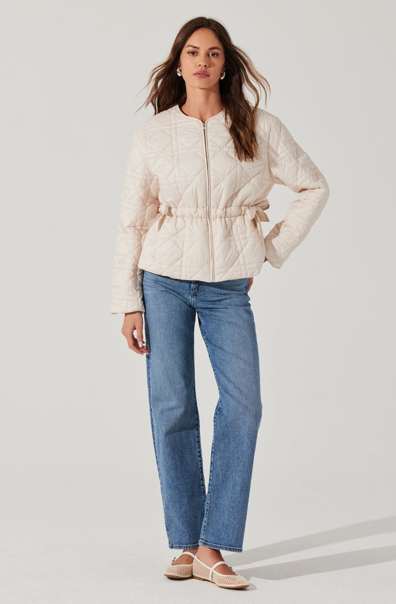 A woman is wearing a light beige quilted jacket with a front zipper, a gathered waist, and long sleeves. She is also wearing straight-leg blue jeans and beige open-toe shoes with a woven design.