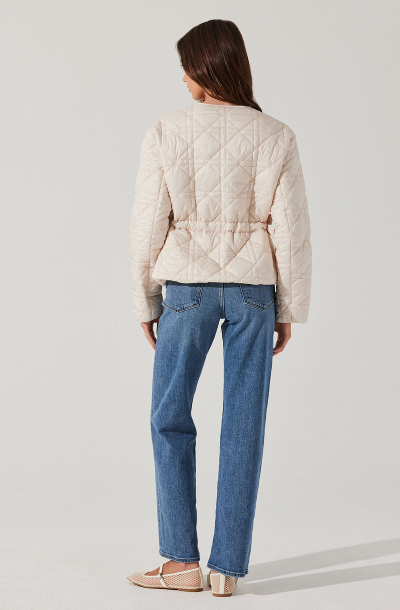 A woman is wearing a light beige, quilted jacket with a fitted waist and diamond-shaped stitching pattern. She pairs it with high-waisted, relaxed-fit blue jeans and beige woven block heel shoes.