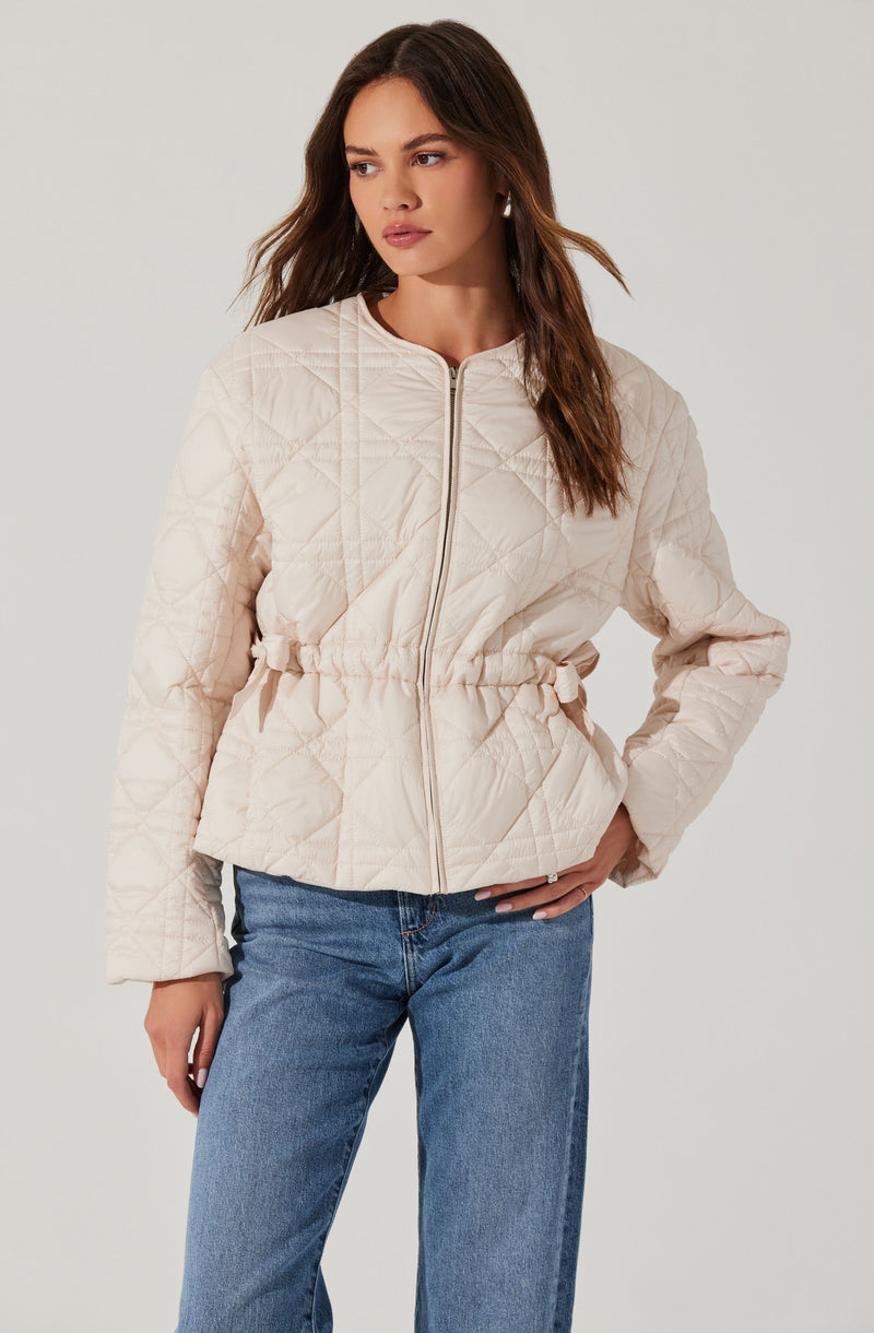 A woman is wearing a light beige quilted jacket with a front zipper, elastic waistband, and long sleeves. The jacket has a diamond quilt pattern and a slightly shiny finish. She pairs it with blue jeans.