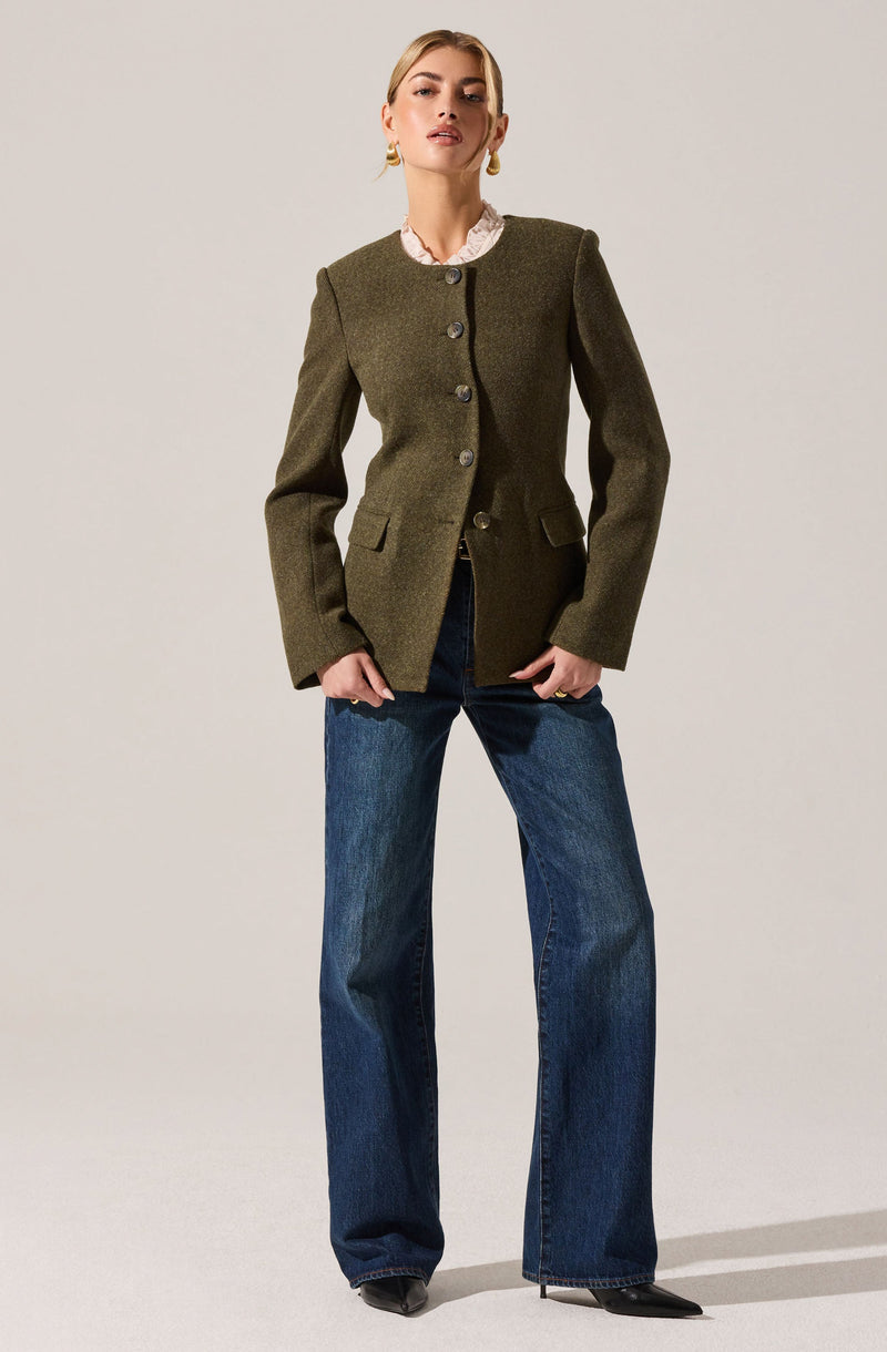 A woman is wearing an olive green tailored blazer with a high neckline, four large buttons down the front, structured shoulders, and flap pockets on each side. She pairs it with wide-leg blue jeans and black pointed shoes. She also has gold hoop earrings and a pink lace collar visible at the neck.