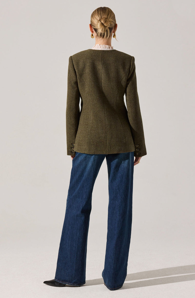 A woman is wearing a tailored, dark green blazer with structured shoulders and long sleeves featuring button details at the cuffs. She has a high-neck, ruffled blouse underneath, which is slightly visible at her neck. She is also wearing wide-leg blue jeans with a high waist, and black shoes peeking out from beneath the hem. Her hair is styled in an elegant updo, and she is accessorized with gold hoop earrings.