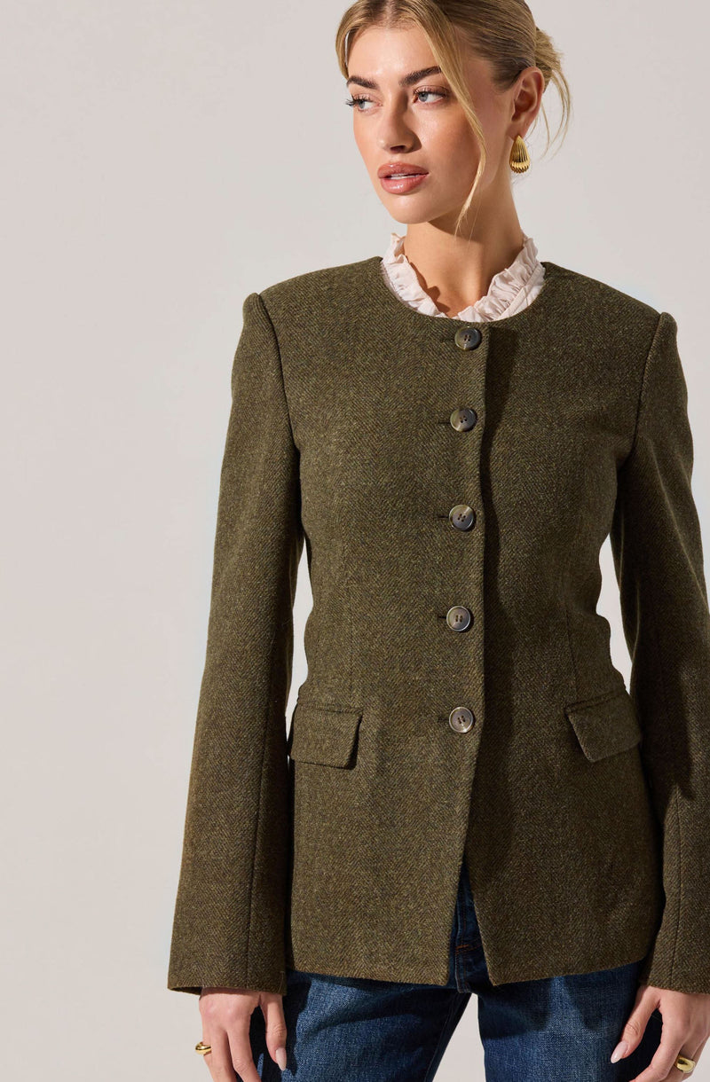 A woman is wearing a dark green, tailored blazer with a textured fabric. The blazer features a round neckline, long sleeves, and a row of large buttons down the front. It also has two flap pockets on the front. Underneath, she is wearing a light-colored, ruffled blouse with a high neckline. She is accessorizing with gold hoop earrings. The woman is dressed in blue jeans.
