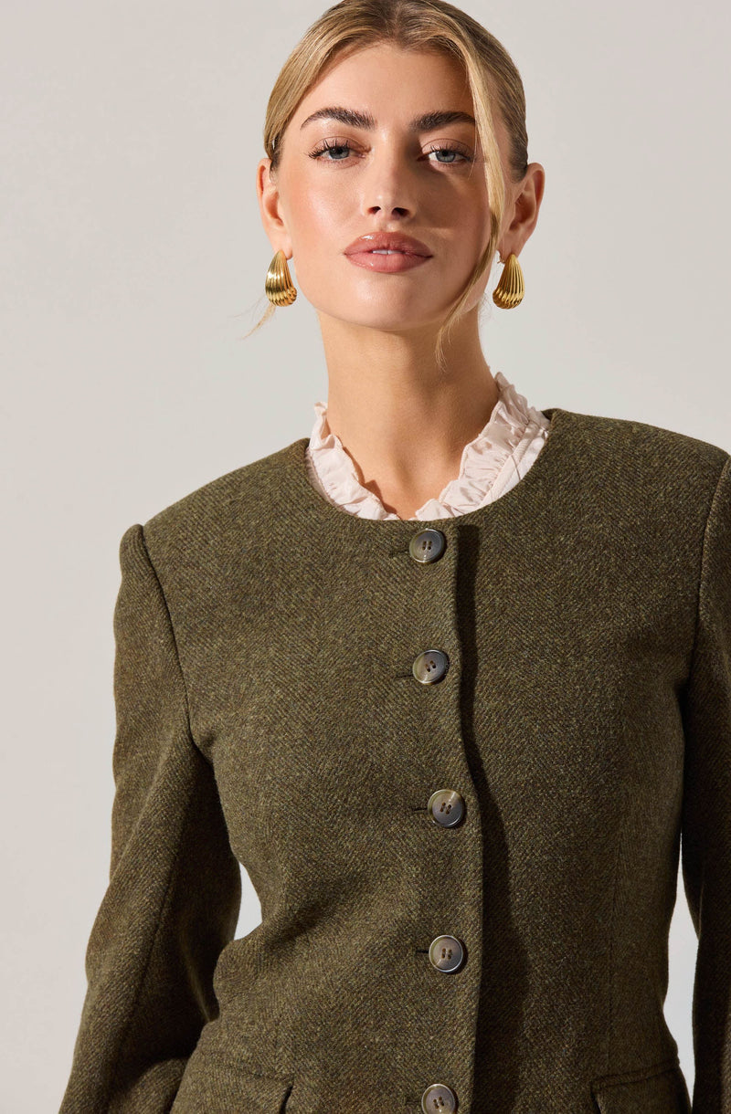A woman is wearing a tailored, olive-green, textured blazer with a round neckline. The blazer features a row of large buttons down the front. She has a white, ruffled blouse underneath, with the ruffles visible at the collar. She is also wearing gold, textured, hoop earrings.