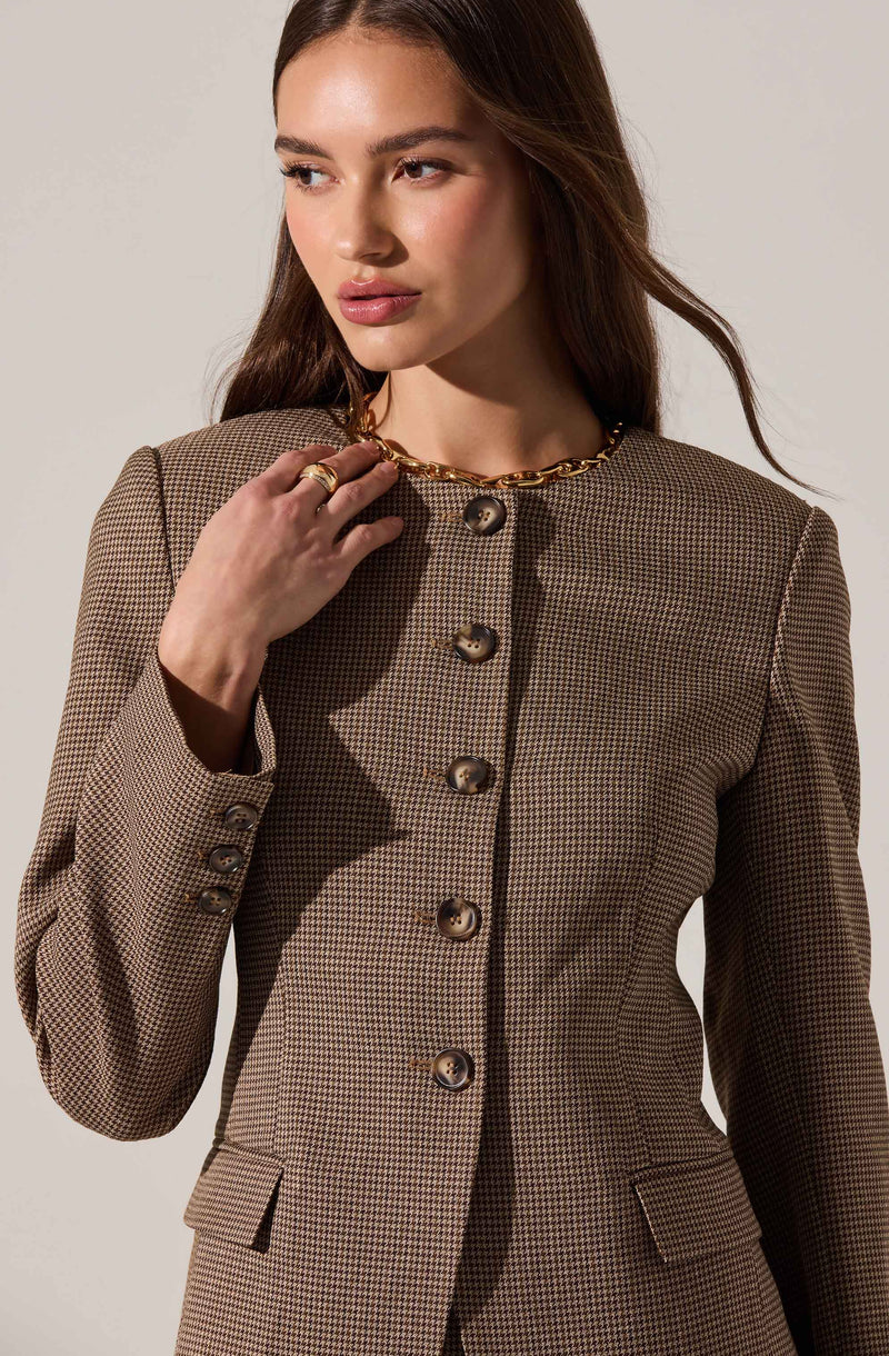 A woman is wearing a tailored, checkered blazer in shades of brown and beige with large, round buttons down the front and a high neckline. She accessorizes with a chunky gold chain necklace and gold rings.