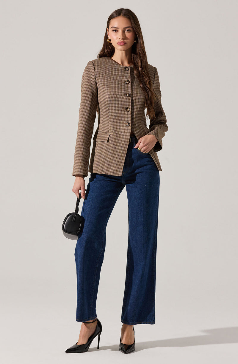 A woman is wearing a tailored, button-up, long-sleeve blazer in a brown houndstooth pattern with a stand-up collar and flap pockets. She pairs it with dark blue wide-leg jeans. She is holding a small black handbag in her left hand and is wearing black high-heeled pumps. She has long, wavy brown hair and is accessorizing with gold hoop earrings.