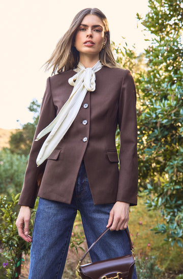 A woman is wearing a tailored brown blazer with a single-breasted design and four buttons down the front. She has a white silk scarf tied around her neck in a bow. She is also wearing high-waisted blue jeans and is holding a dark brown handbag.