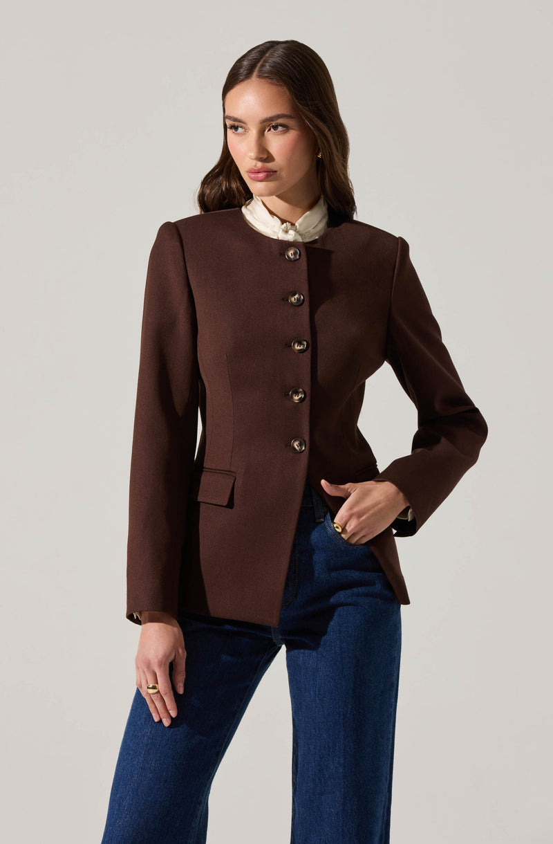 A woman is wearing a fitted, dark brown blazer with structured shoulders and a high neckline. The blazer features a row of large, shiny buttons down the front and has a small pocket flap on the left side. Underneath, she has on a white, ruffled collared blouse. She pairs the blazer with dark blue, straight-leg jeans.