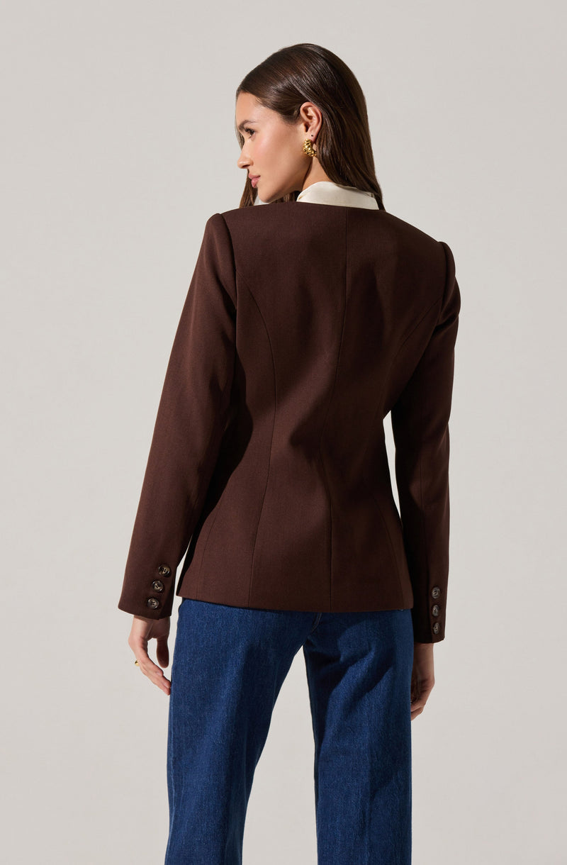A woman is wearing a tailored dark brown blazer with structured seams and button detail on the cuffs. The blazer features a white collar peeking out from underneath. She is also wearing blue jeans.