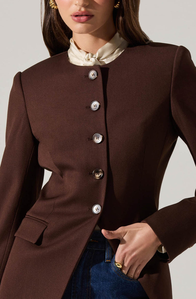 A woman is wearing a fitted, dark brown blazer with a structured shoulder design. The blazer features a row of shiny, light-colored buttons down the front. She is also wearing a white, silk or satin scarf tied around her neck, and gold hoop earrings. Her right hand is in her pocket, revealing a gold ring on her finger.