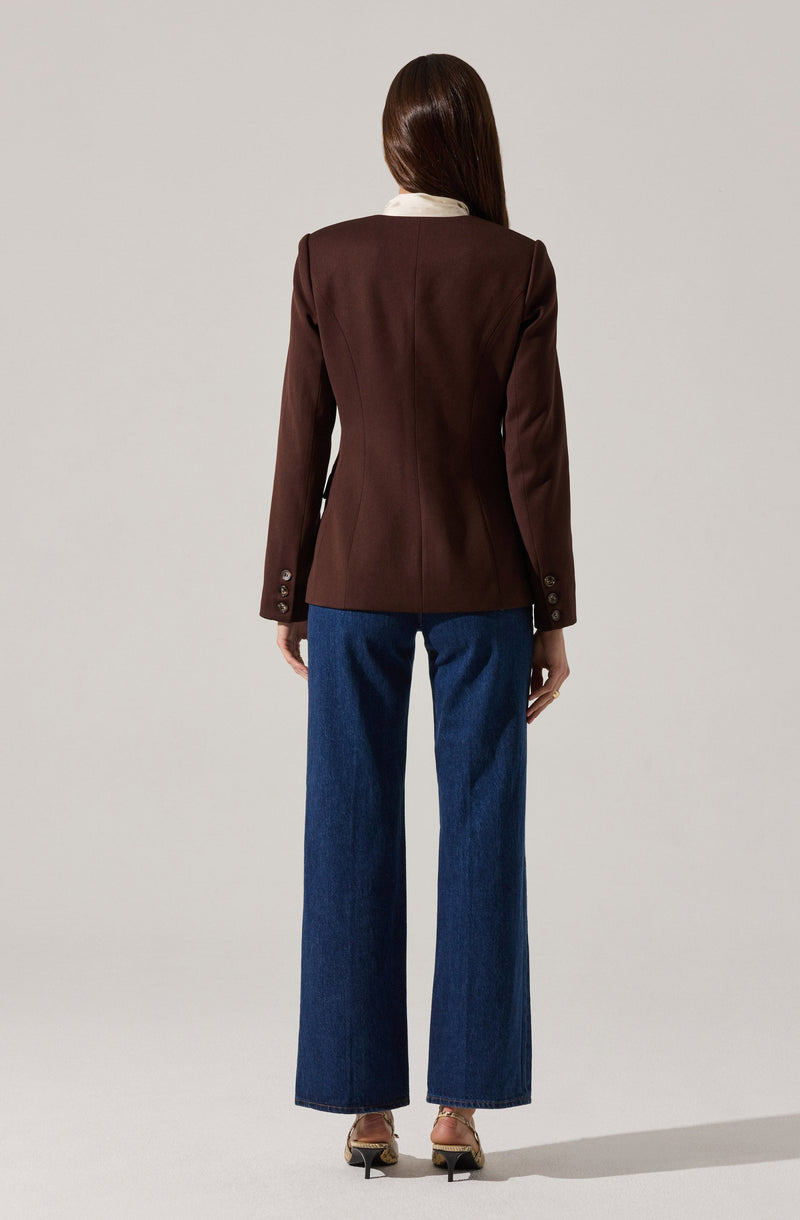 A person wearing a fitted dark brown blazer with long sleeves and button details at the cuffs, paired with wide-leg blue jeans and beige high-heeled shoes.