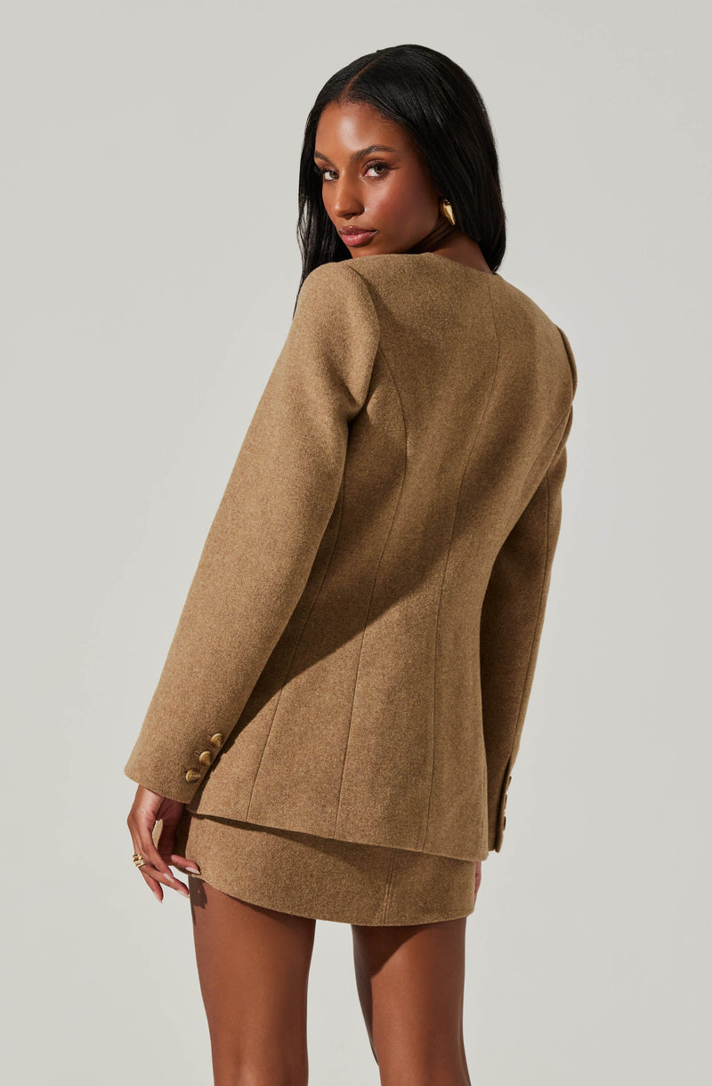 A woman is wearing a tan-colored, tailored blazer with a matching mini skirt. The blazer features a structured fit, with seam details and gold buttons on the sleeves. The outfit appears to be made of a wool or wool-blend fabric.
